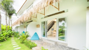A modern building with a thatched roof, a staircase leading to a glass door, and a well-maintained garden with stepping stones and tropical plants. - Baan Seanok lofts (Pecatu)