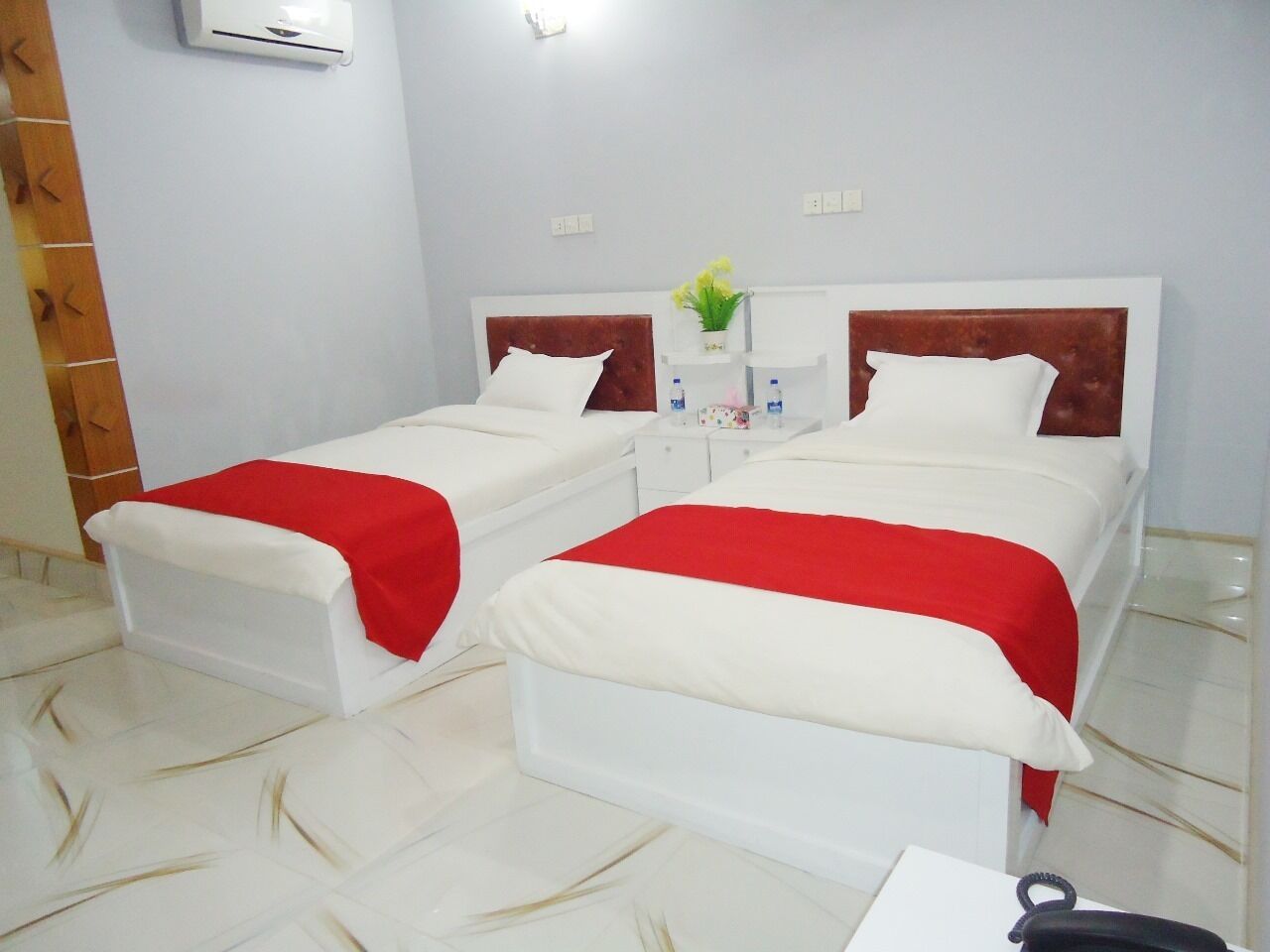 A hotel room with two beds, each with a red and white bedspread, white headboards, and a small bedside table with a vase of flowers.