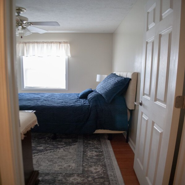 A bedroom with a bed, a window with curtains, a ceiling fan, and a door.