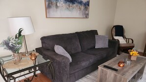 A living room with a dark grey sofa, a wooden coffee table, a glass table with a lamp, a vase with flowers, and a painting on the wall. - Quiet and sophisticated apartment with AC in Clinton (Clinton)