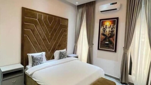 Une chambre moderne avec un grand lit, une tête de lit ornée d’un motif géométrique, une table de chevet, une commode, un banc, un appareil de climatisation et un tableau encadré accroché au mur.