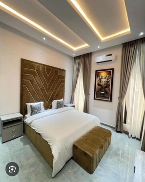 Une chambre moderne avec un grand lit, une tête de lit ornée d’un motif géométrique, une table de chevet, une commode, un banc, un appareil de climatisation et un tableau encadré accroché au mur.