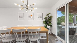 A dining area with a wooden table, white chairs, a modern chandelier, and framed artwork on the wall.