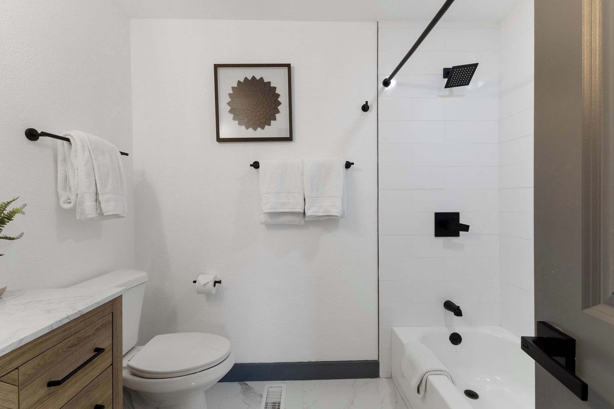 A modern bathroom with a toilet, sink, and bathtub. There are towels hanging on hooks and a framed picture on the wall.
