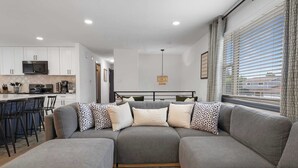 A modern living room with a grey sectional sofa, a dining area with bar stools, and a kitchen with white cabinets and a microwave.