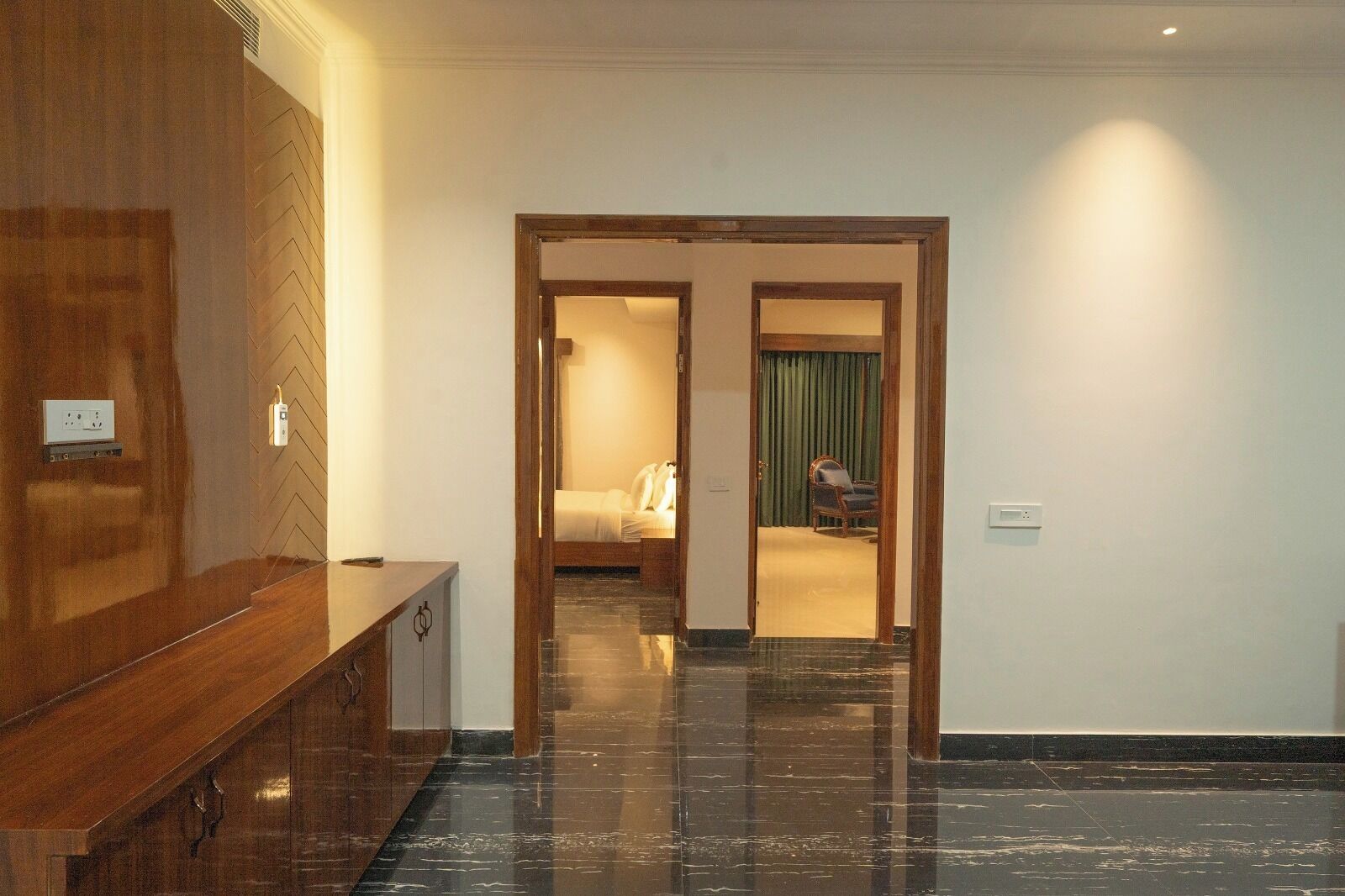 A hotel hallway with a wooden reception desk, a bed in the background, and a chair by the window.
