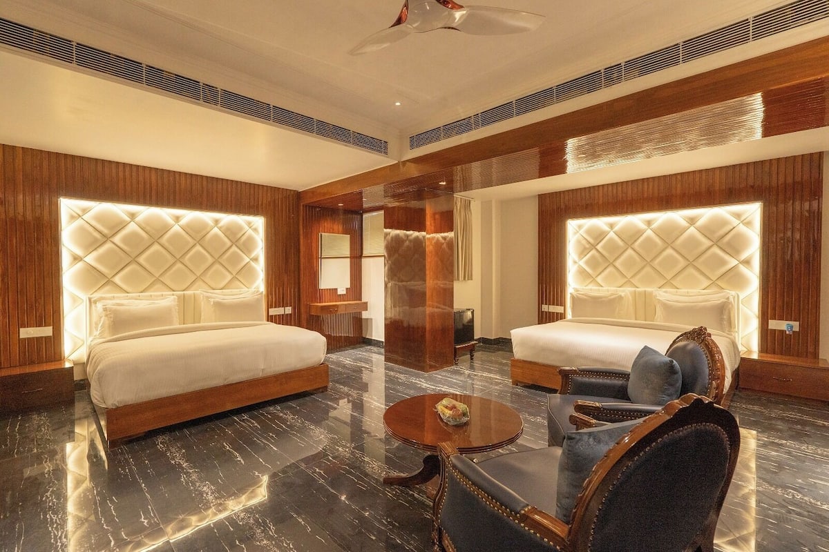 Quarto de hotel luxuoso com duas camas grandes, um espaço de estar com mesa de centro e um espelho amplo.