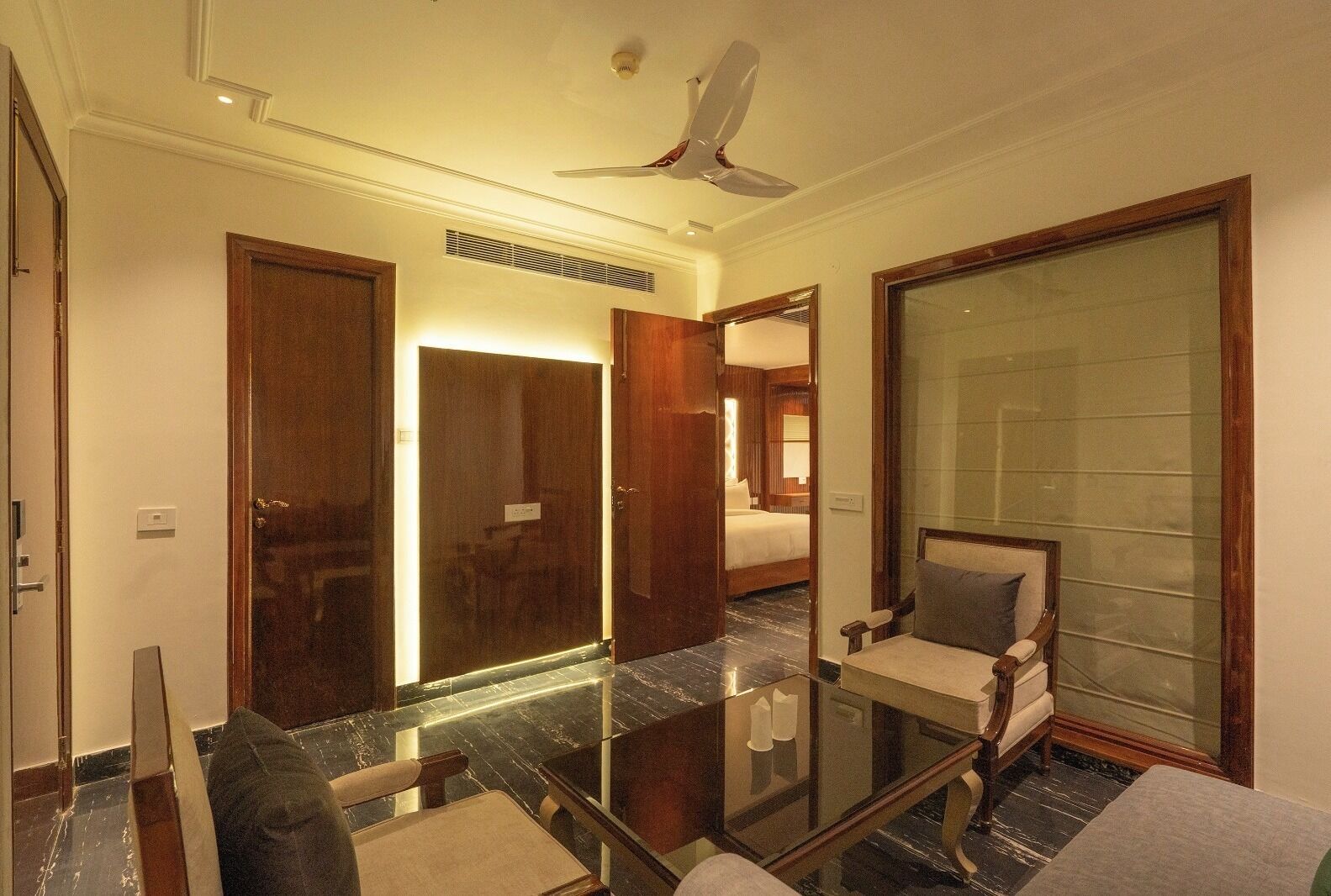 Habitación de hotel con una mesa de vidrio, una silla, una cama, un espejo grande y un ventilador de techo.