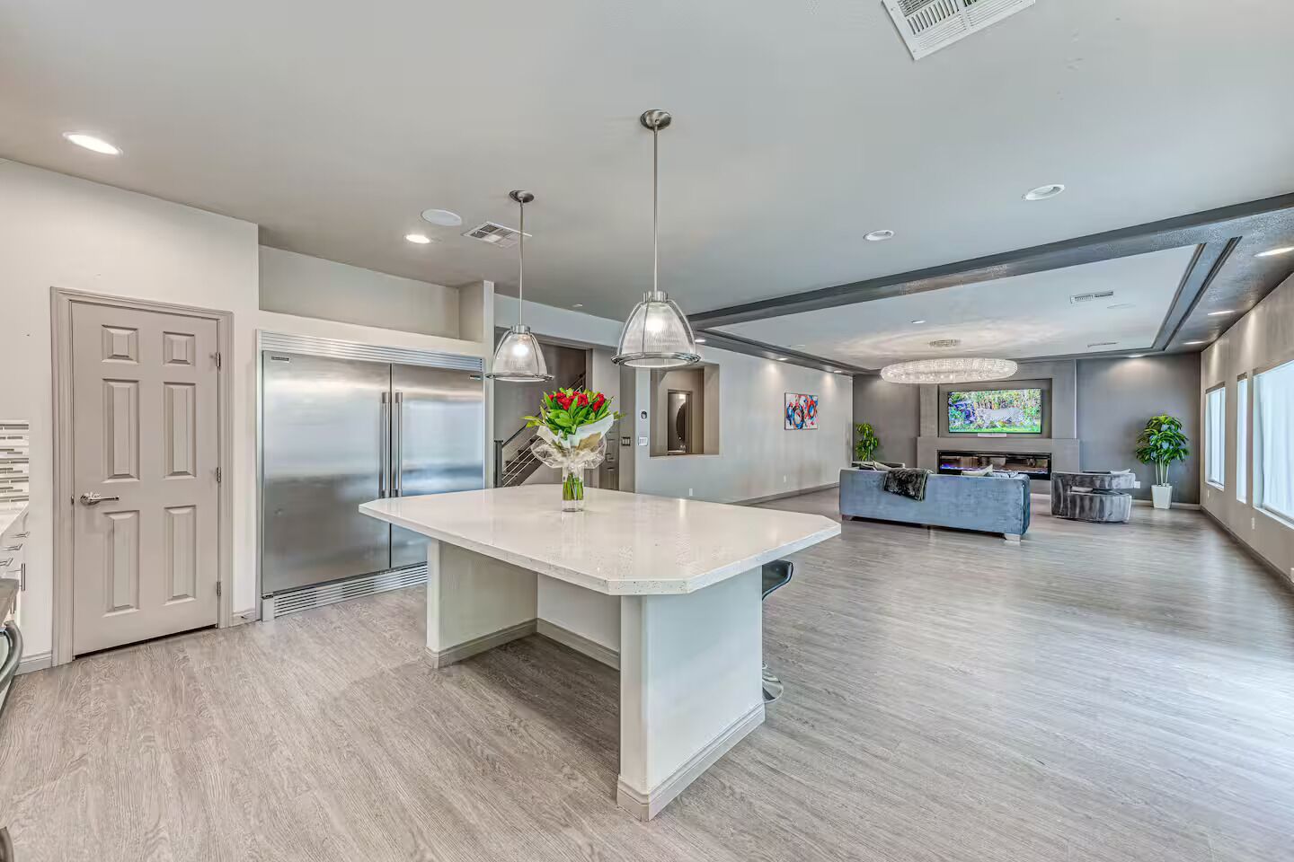 A modern kitchen with a central island, stainless steel appliances, pendant lighting, and a large TV in the background.