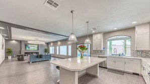 A modern kitchen with white cabinets, a large island, and a dining area with a gray sofa and TV. - Luxury 2 Story 3 Car Garage Home (Las Vegas)