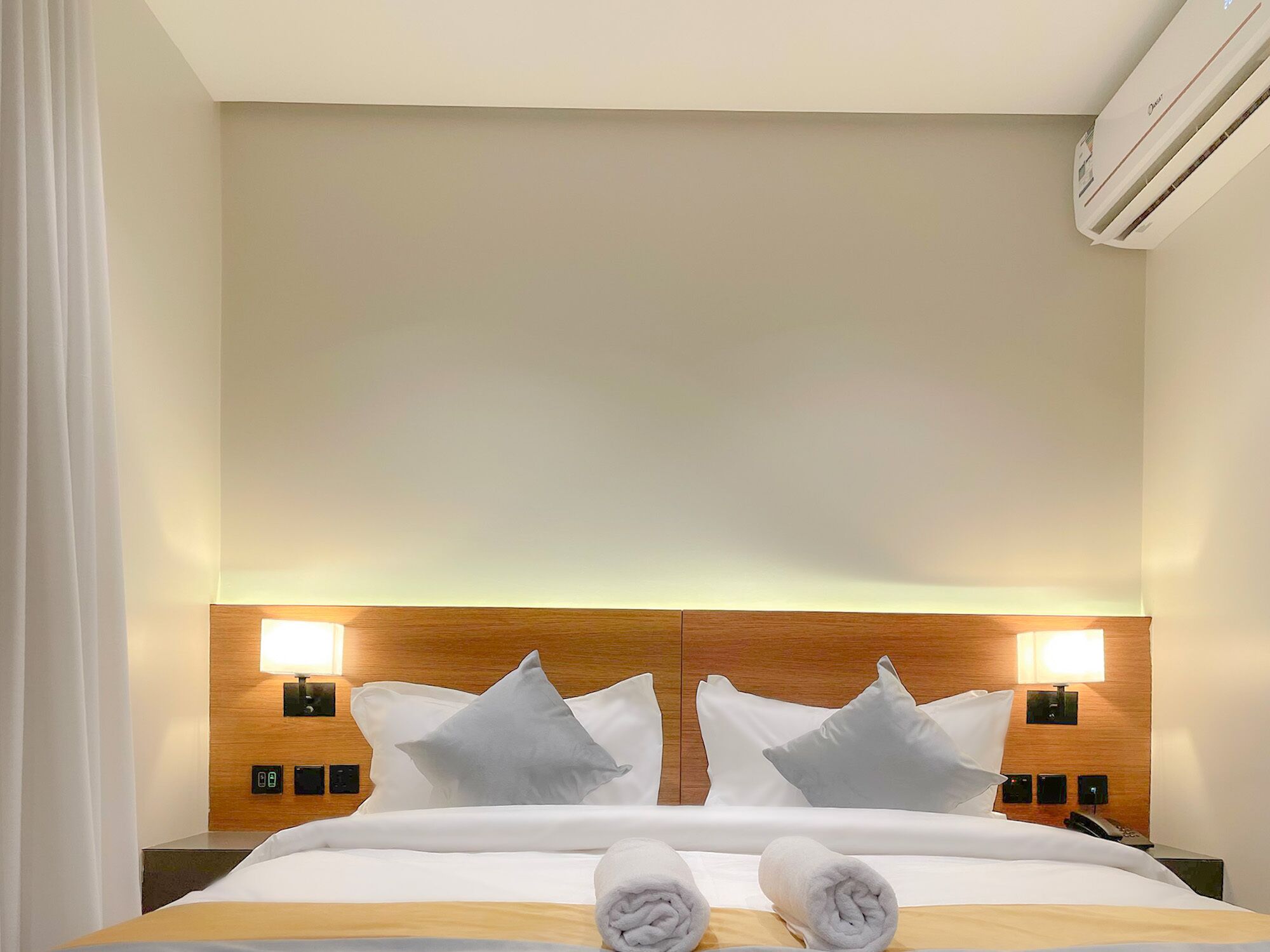 A hotel room with a large bed, two bedside lamps, a wooden headboard, and a wall-mounted air conditioner.