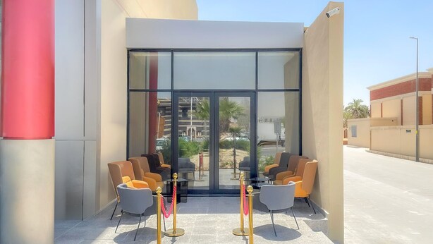 A hotel entrance with a glass door, a seating area with chairs, and a security barrier. - Arjan Park Apartments (Riyadh)