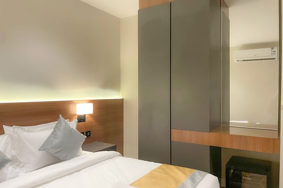 A hotel room with a bed, pillows, a bedside table, a lamp, a wardrobe, and an air conditioning unit.