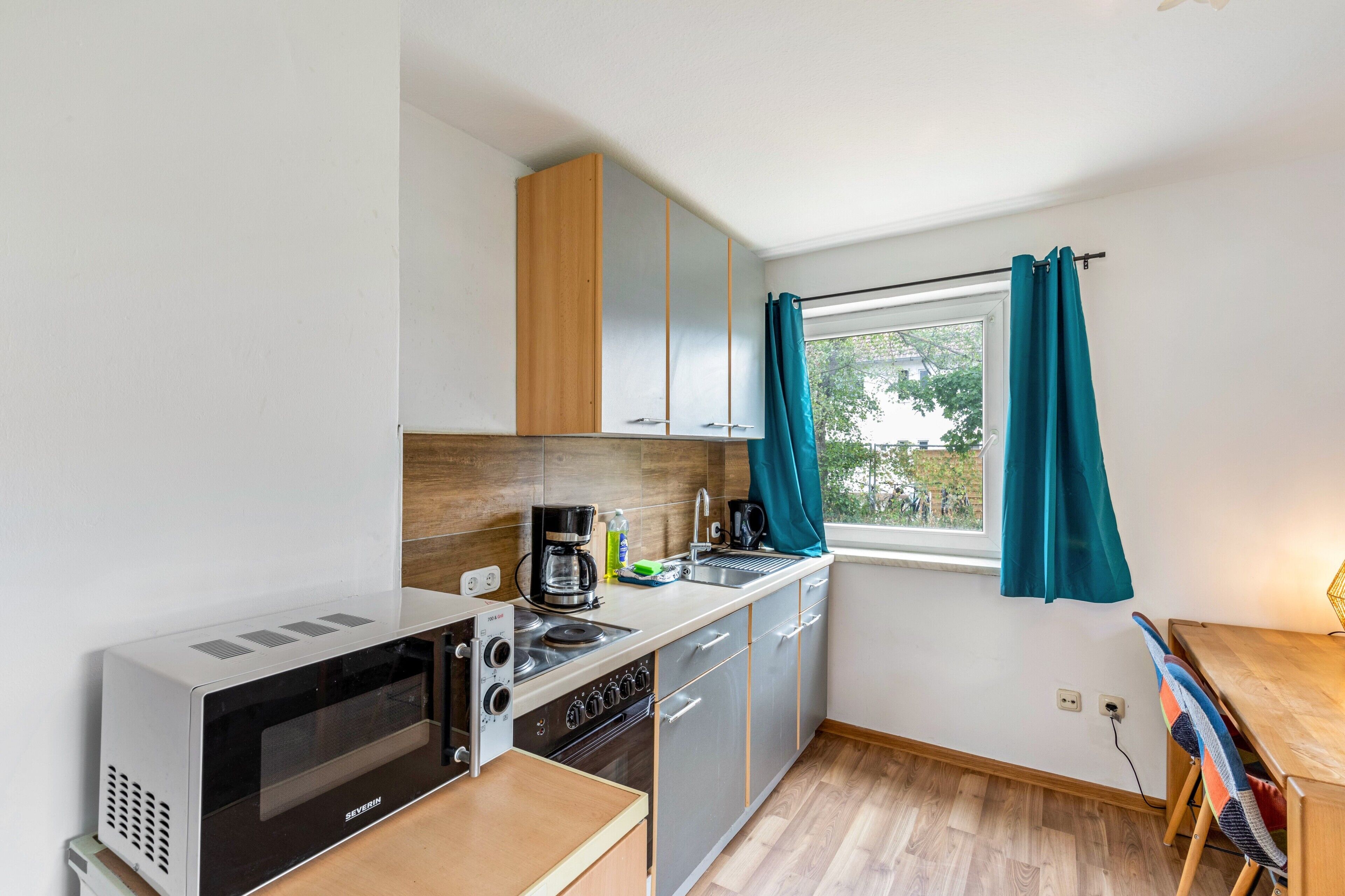 A compact kitchen with a microwave, stove, and sink, wooden cabinets, and a window with curtains.