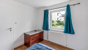 A bedroom with a wooden dresser, a window with teal curtains, a radiator, and a view of buildings outside. - Apartment 'Yellow A' (Miltenberg)