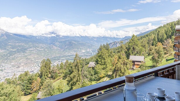 Vista de montanha a partir de uma varanda com uma mesa posta para o chá, com vista para um vale com edifícios e uma estrada.