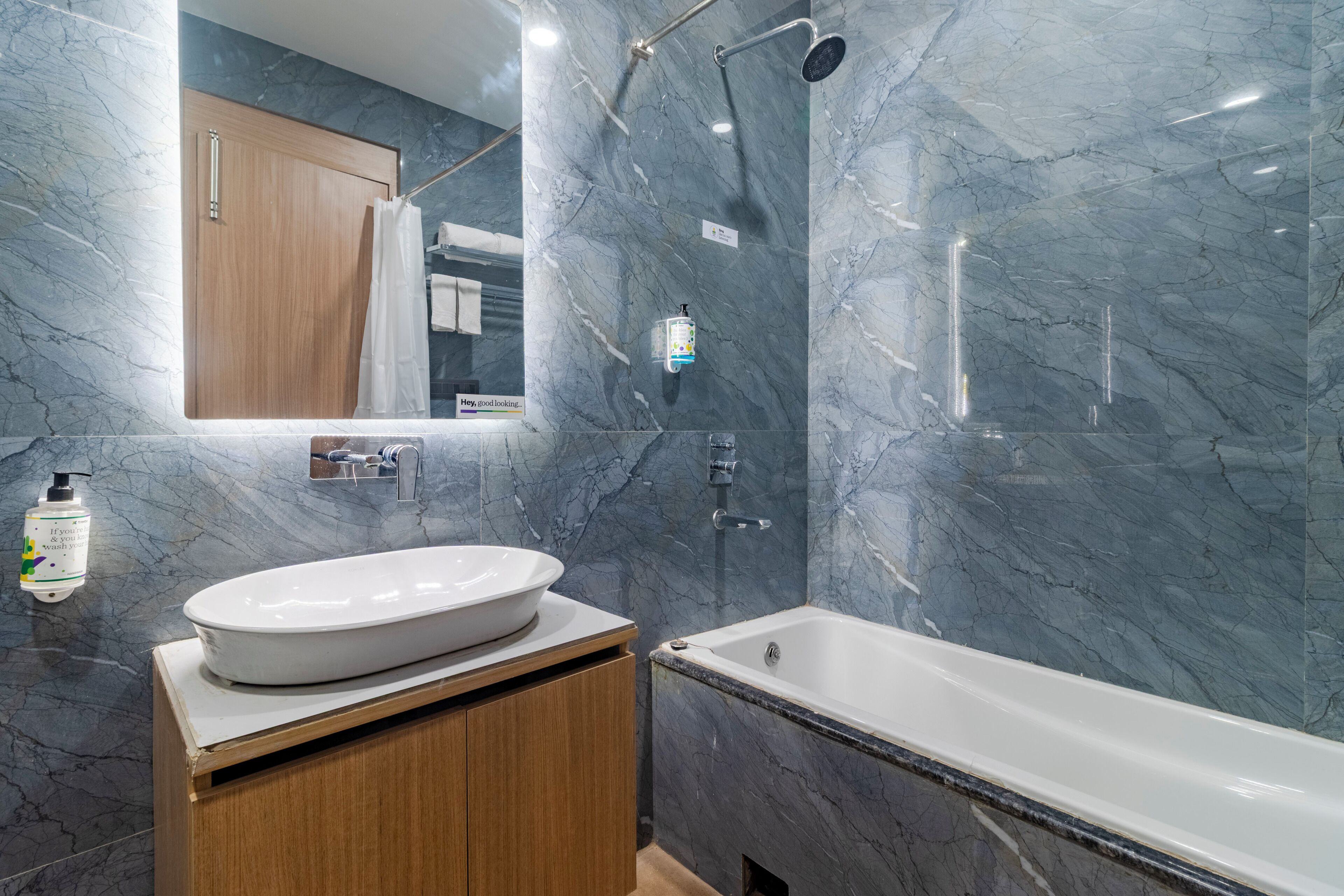 A modern bathroom with a marble wall, a wooden vanity, a white sink, a bathtub, and a shower with a handheld showerhead.