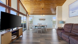 A spacious living area with a large flat-screen TV, wooden flooring, a dining table with chairs, and a brown leather sofa.