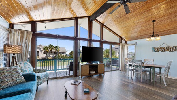 A spacious living room with a wooden ceiling, large windows, a flat-screen TV, a coffee table, and a dining area with chairs and a table.