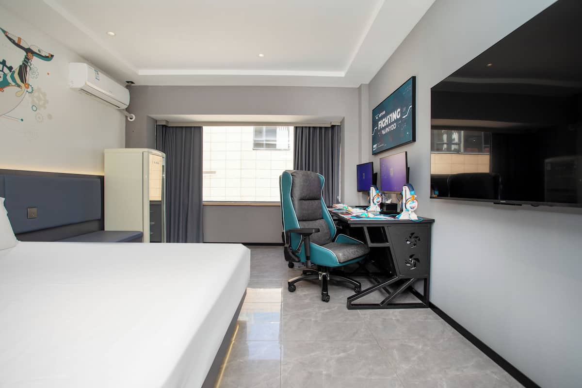 Ein Hotelzimmer mit einem Bett, einem Schreibtisch mit Computer, einem Stuhl, einer Klimaanlage, einem Fernseher und einem Fenster mit Vorhängen.
