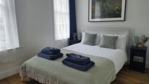 Een netjes opgemaakt bed met een geborreld sprei en opgevouwen handdoeken, een ingelijst kunstwerk erboven, en een nachtkastje met een lamp en een kleine vaas.