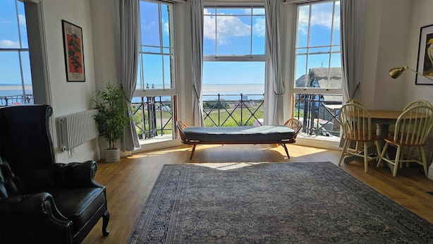 Een kamer met een groot erkerraam, een zwarte leren fauteuil, een houten tafel met twee stoelen, een kamerkleed met patroon en uitzicht op zee.