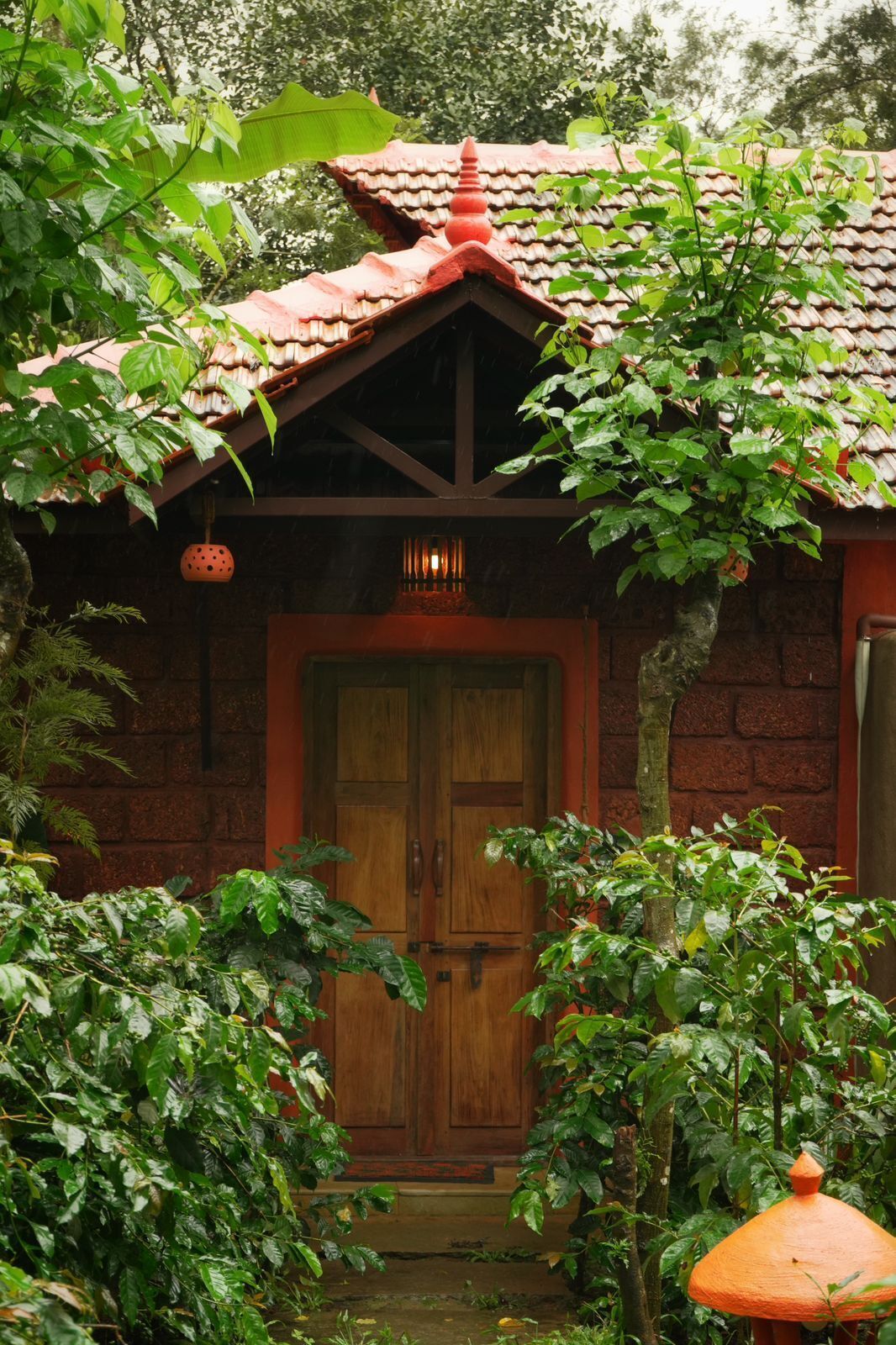 A traditional house with a tiled roof, a wooden door, and a small porch area surrounded by lush greenery.