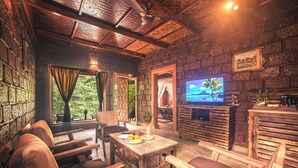 A cozy room with a stone wall, wooden furniture, a television, and a view of greenery outside. -  Copper Edge – Eco Resort, Coorg  (Shanivarsanthe)
