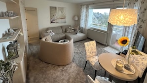 A cozy living room with a sectional sofa, a round coffee table, and a dining area with chairs. There is a large window with curtains, a painting on the wall, and a lamp on the table. - Modern, cozy, close to the beach - vacation apartment in Cuxhaven, Döse, right on the beach (Cuxhaven)