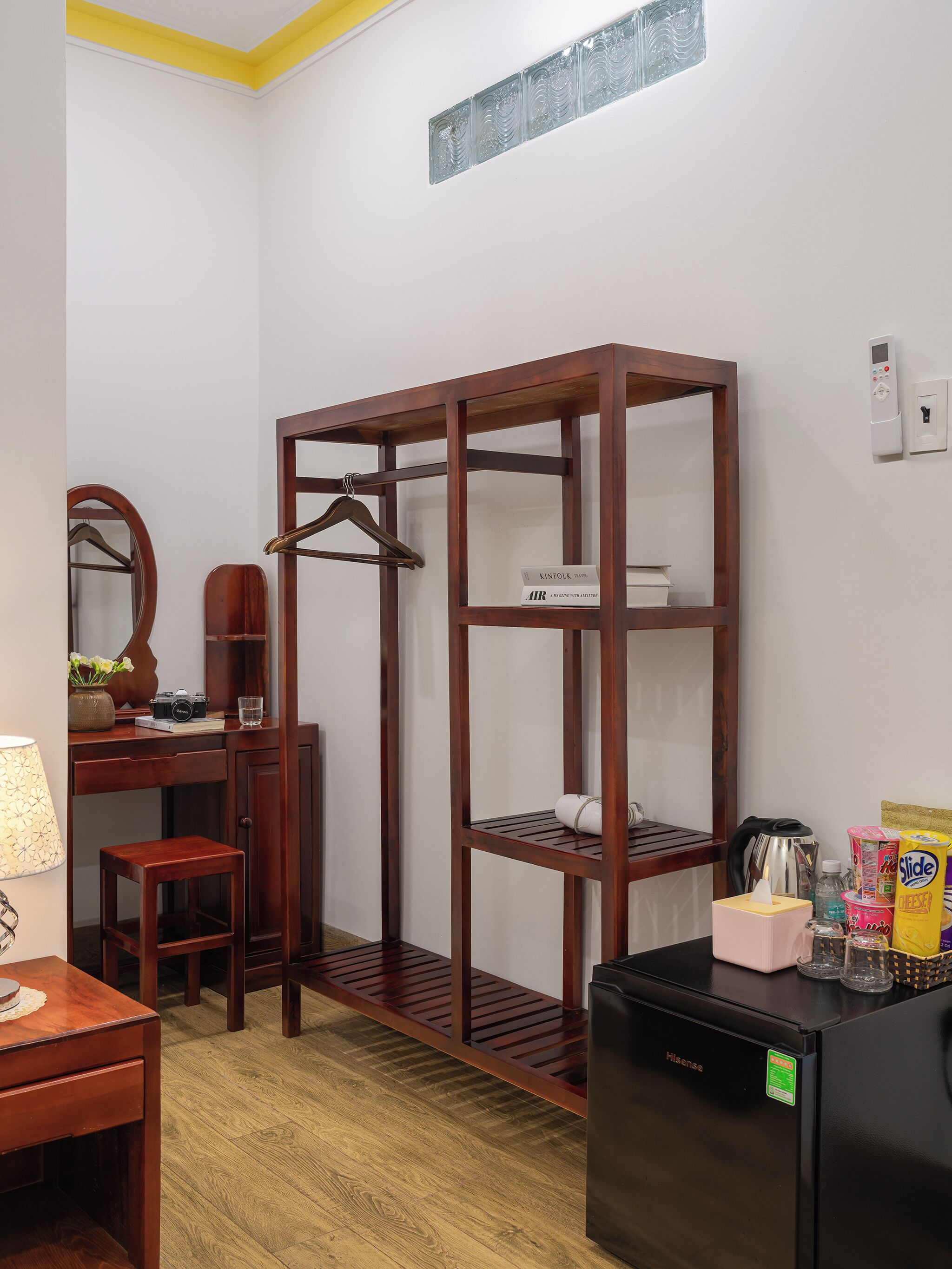 A compact room with a wooden wardrobe, a desk with a mirror, a chair, a small table, and a refrigerator.