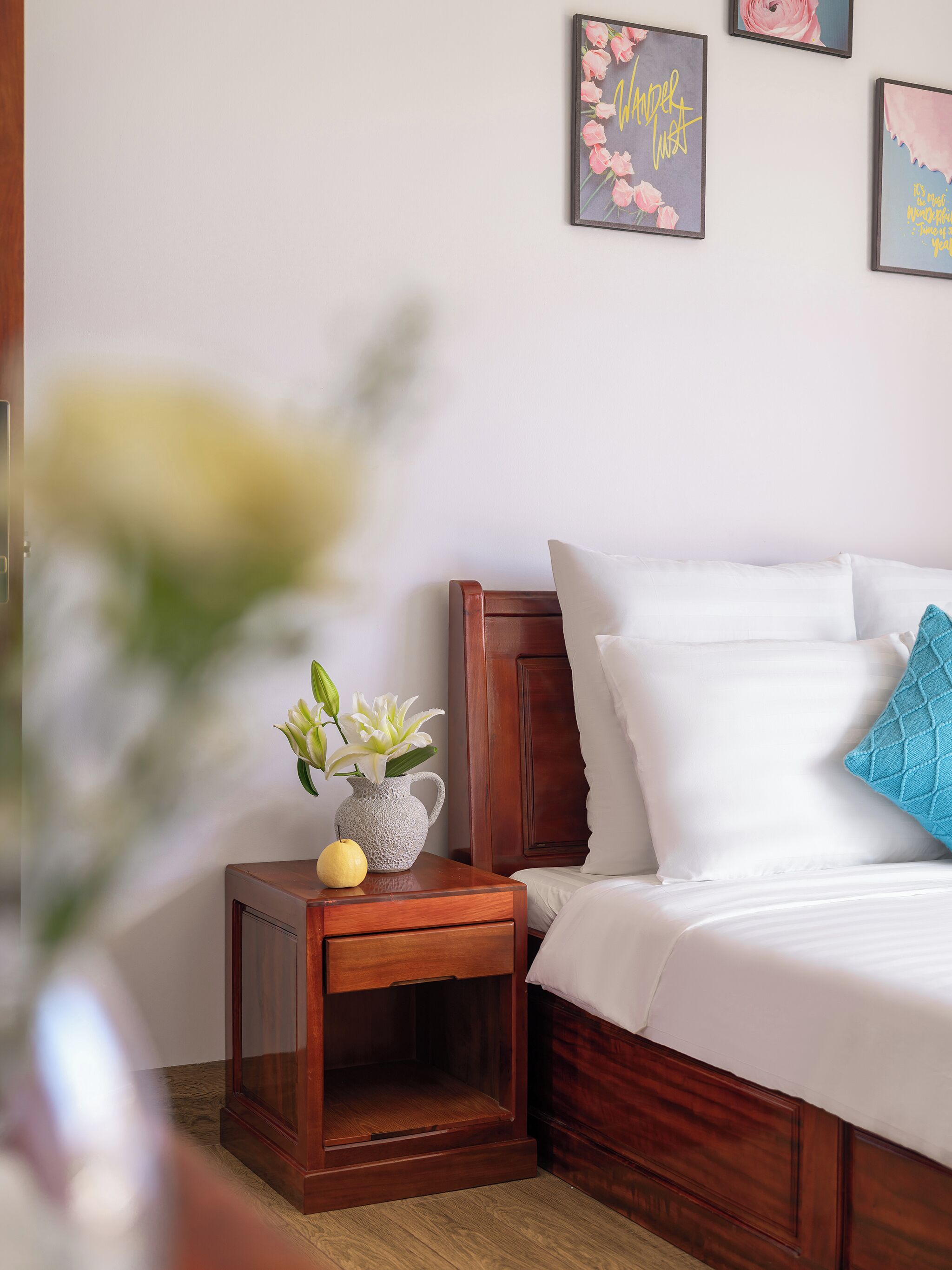 A neatly made bed with white linens, a wooden nightstand with a vase of flowers, and three framed pictures on the wall.