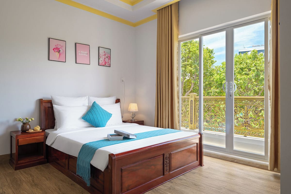 A neatly made bed with a blue pillow, a bedside table with a lamp, and a window with a view of greenery and a building.