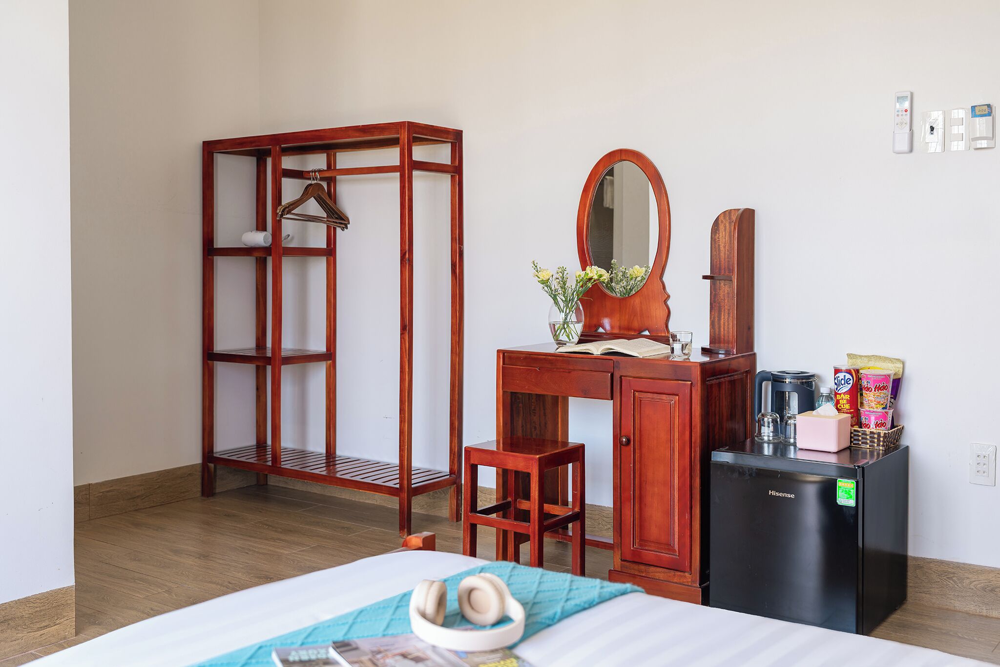 A bedroom with a bed, a wooden wardrobe, a desk with a mirror, a small stool, and a black refrigerator.