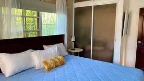Un dormitorio con cama, almohadas blancas, colcha azul, una mesita con lámpara, una puerta corredera y una ventana con cortinas.