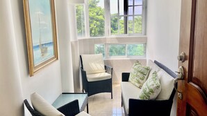 A sunlit room with a view of greenery, a wicker chair with a cushion, a sofa with patterned cushions, and a framed painting on the wall.