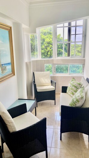 Una habitación iluminada por el sol con muebles de mimbre, un cuadro enmarcado y vistas a la vegetación a través de amplias ventanas.