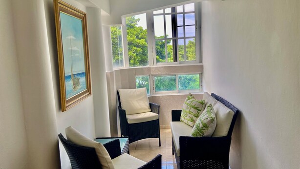 A sunlit room with a view of greenery through large windows, a framed painting, and a seating area with wicker furniture.
