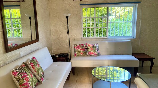 Una sala de estar con un sofá blanco, una mesa redonda de cristal y un ventanal con vistas a zonas verdes.