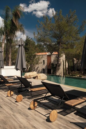A poolside area with lounge chairs and umbrellas, surrounded by trees and a stone building.