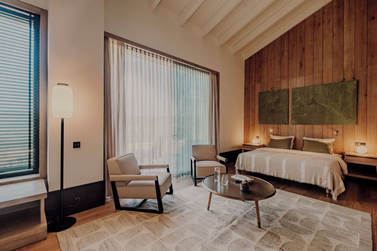 A modern hotel room with a large bed, two armchairs, a coffee table, and a large window with blinds.