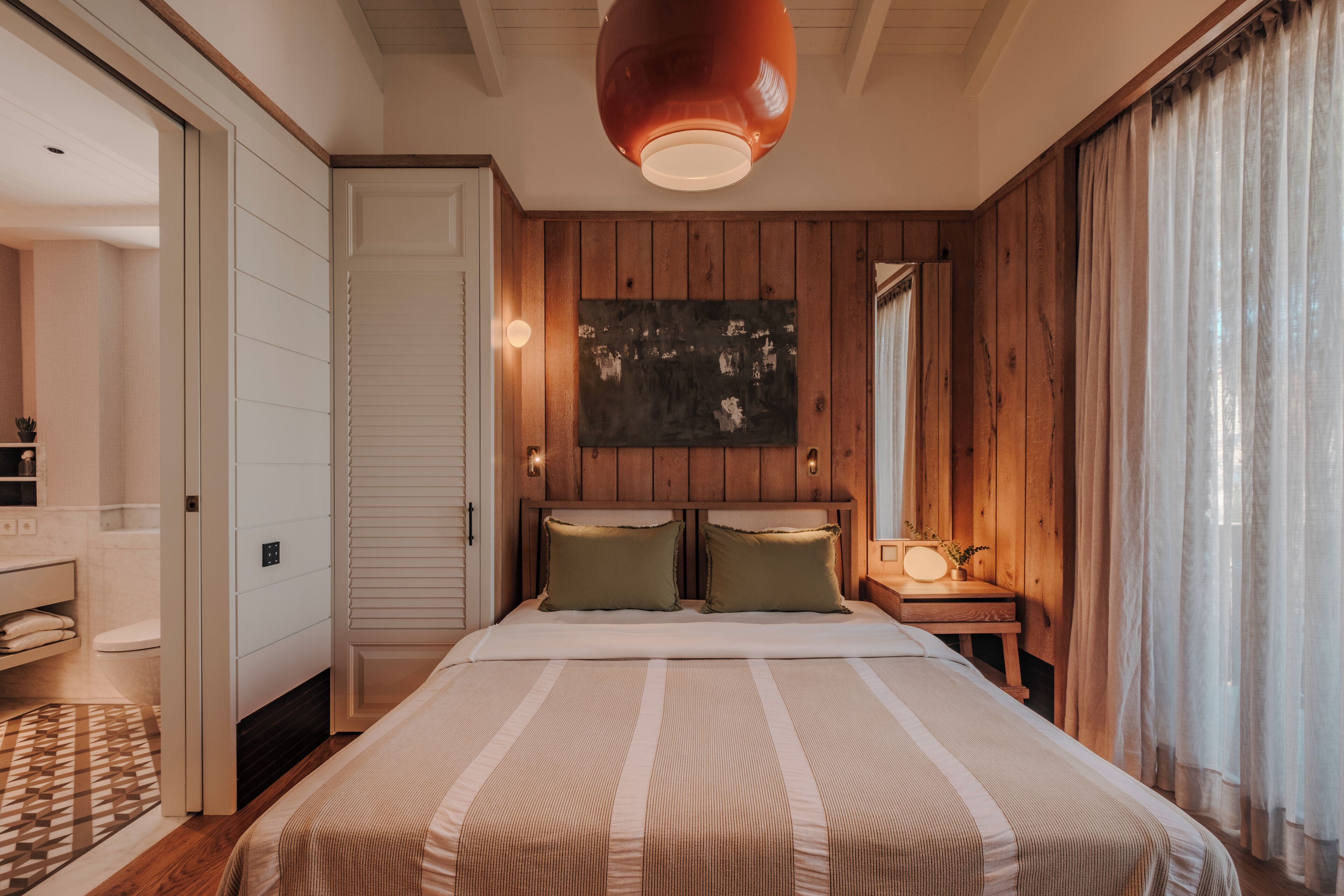 A bedroom with a large bed, wooden panel walls, a bedside table with a lamp, and a painting on the wall.