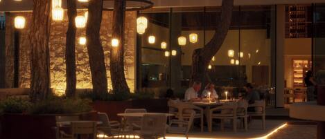 Un espacio de comedor al aire libre con mesas y sillas, rodeado de árboles e iluminado por luces colgantes.