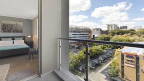 Una habitación de hotel moderna con vista a la ciudad, un balcón con barandilla de vidrio y una cama bien hecha.