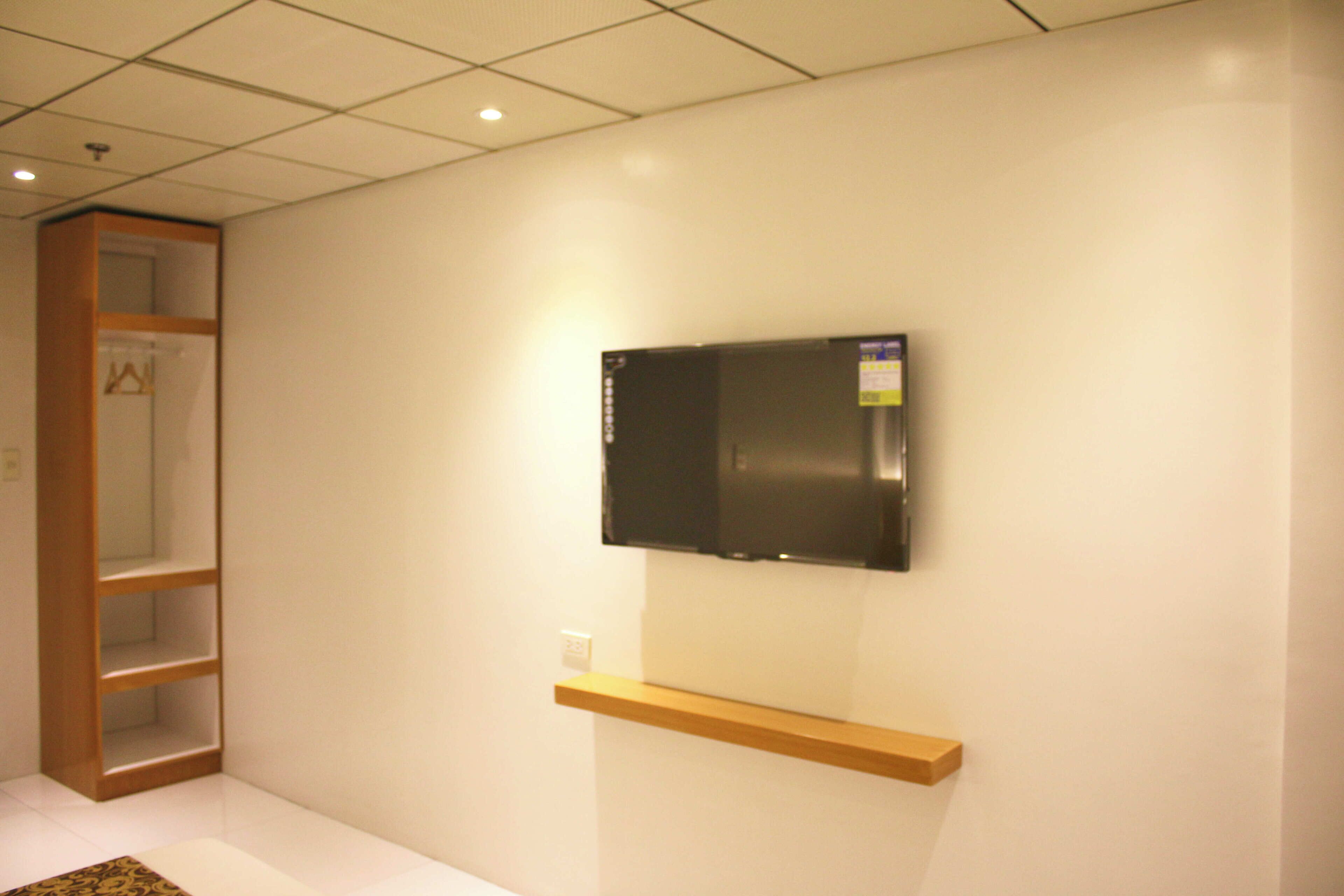 A room with a mounted flat-screen TV on a wooden shelf, a built-in wooden shelving unit, and a white tiled floor.