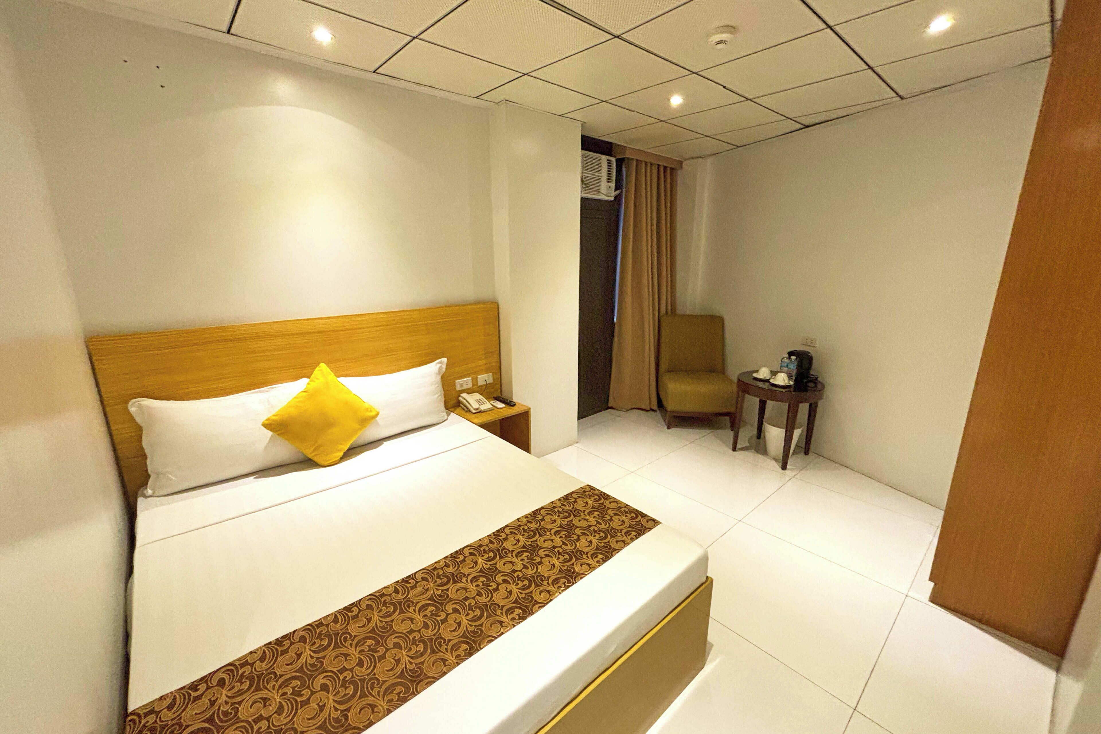 A hotel room with a bed, a bedside table with a phone, a chair, and a small table with a cup and saucer.