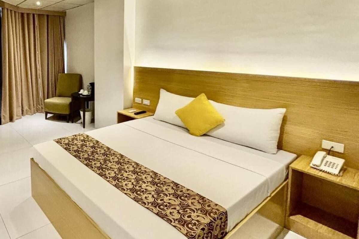 A hotel room with a large bed, a bedside table with a phone, a chair, and a desk with a lamp.