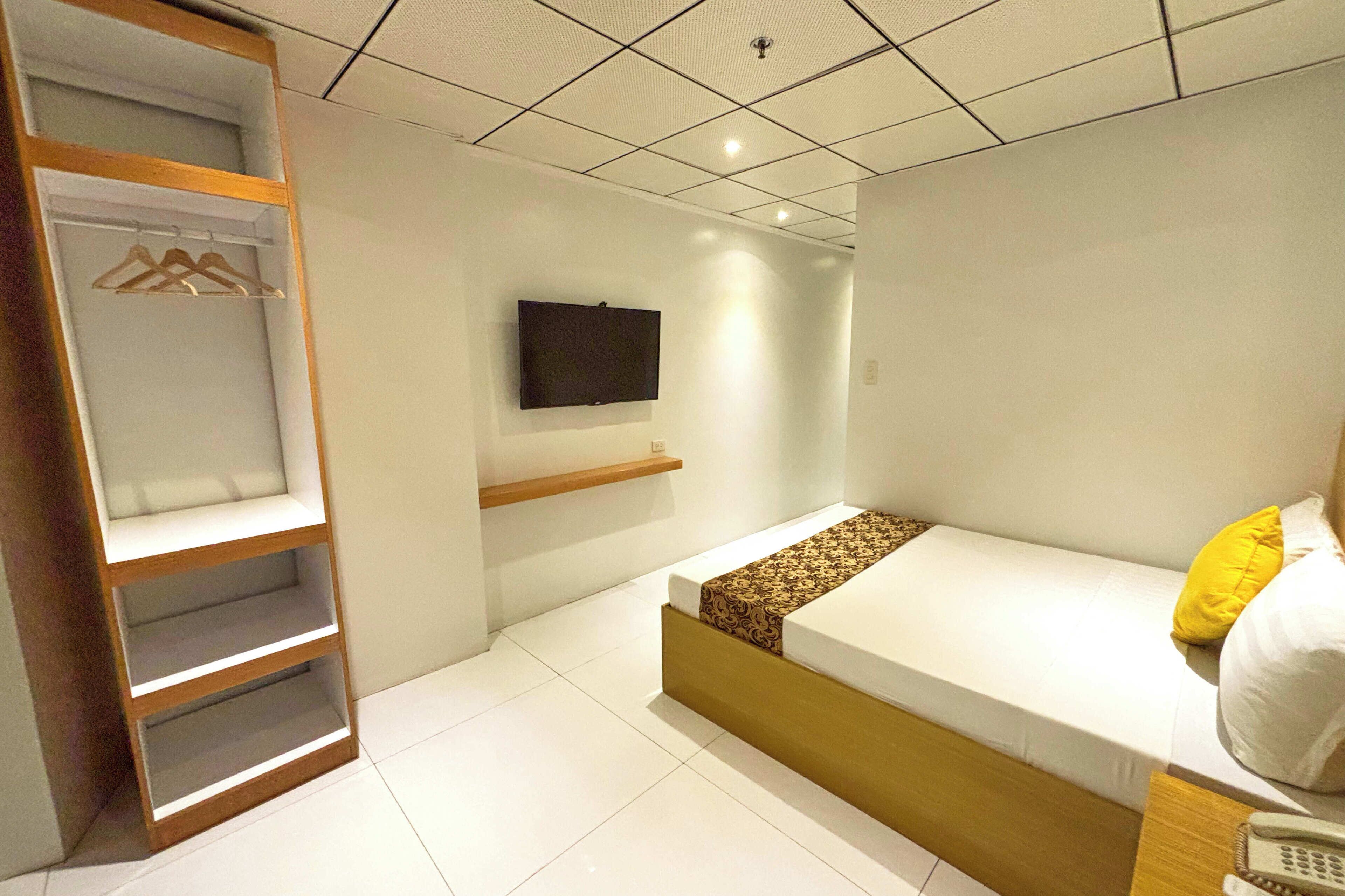 A bedroom with a bed, a TV mounted on the wall, a wooden wardrobe, and a shelf.