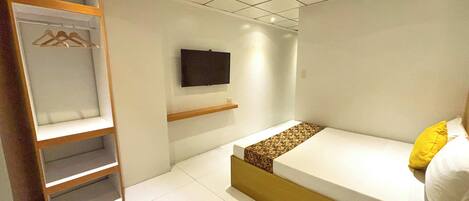 A bedroom with a bed, a TV mounted on the wall, a wooden wardrobe, and a shelf.