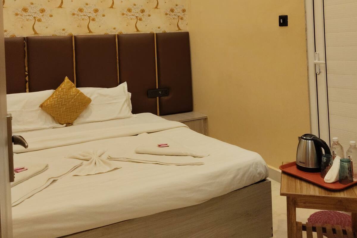 A hotel room with a bed, a nightstand, a table with a kettle, and a chair.