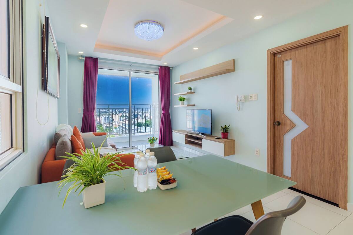 Ein modernes Wohnzimmer mit einem Glastisch, einer Couch mit orangefarbenen Kissen, einem Fernseher auf einem Holzboden und einem Balkon mit violetten Vorhängen.
